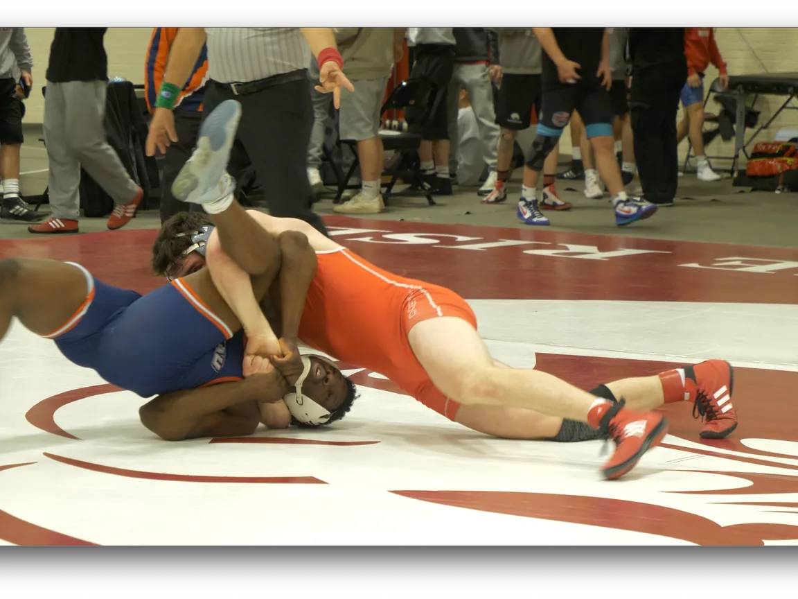 Two high school wrestlers in singlets battling for control during a dual meet, with teammates lining the sideline.