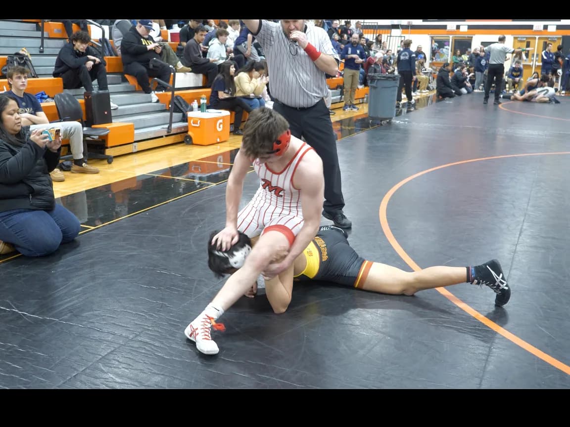 A wrestler controls his opponent on the mat while a parent watches from the front-row bleachers.