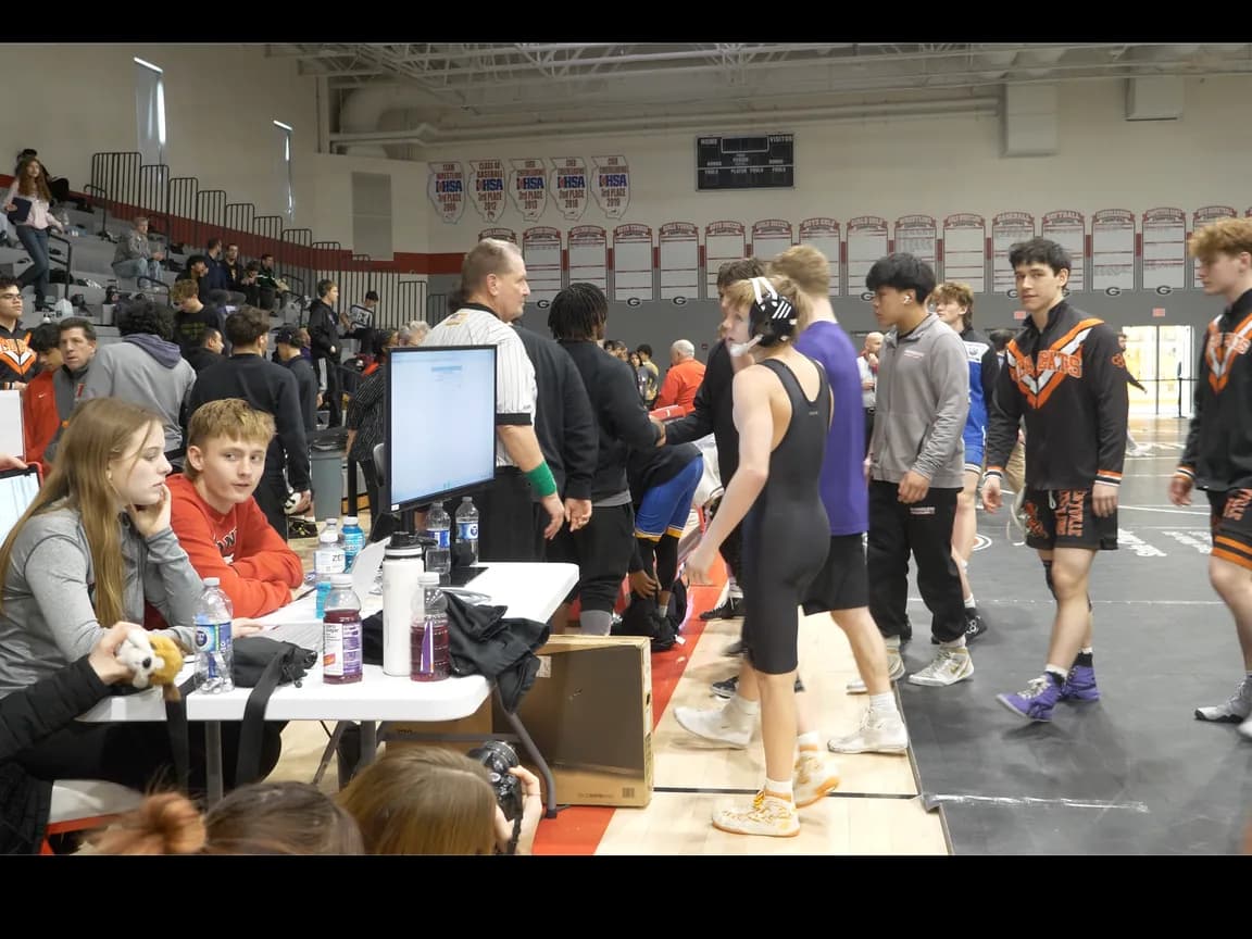 A scoring table beside the mat with a referee and athletes preparing for the next bout at a wrestling tournament.