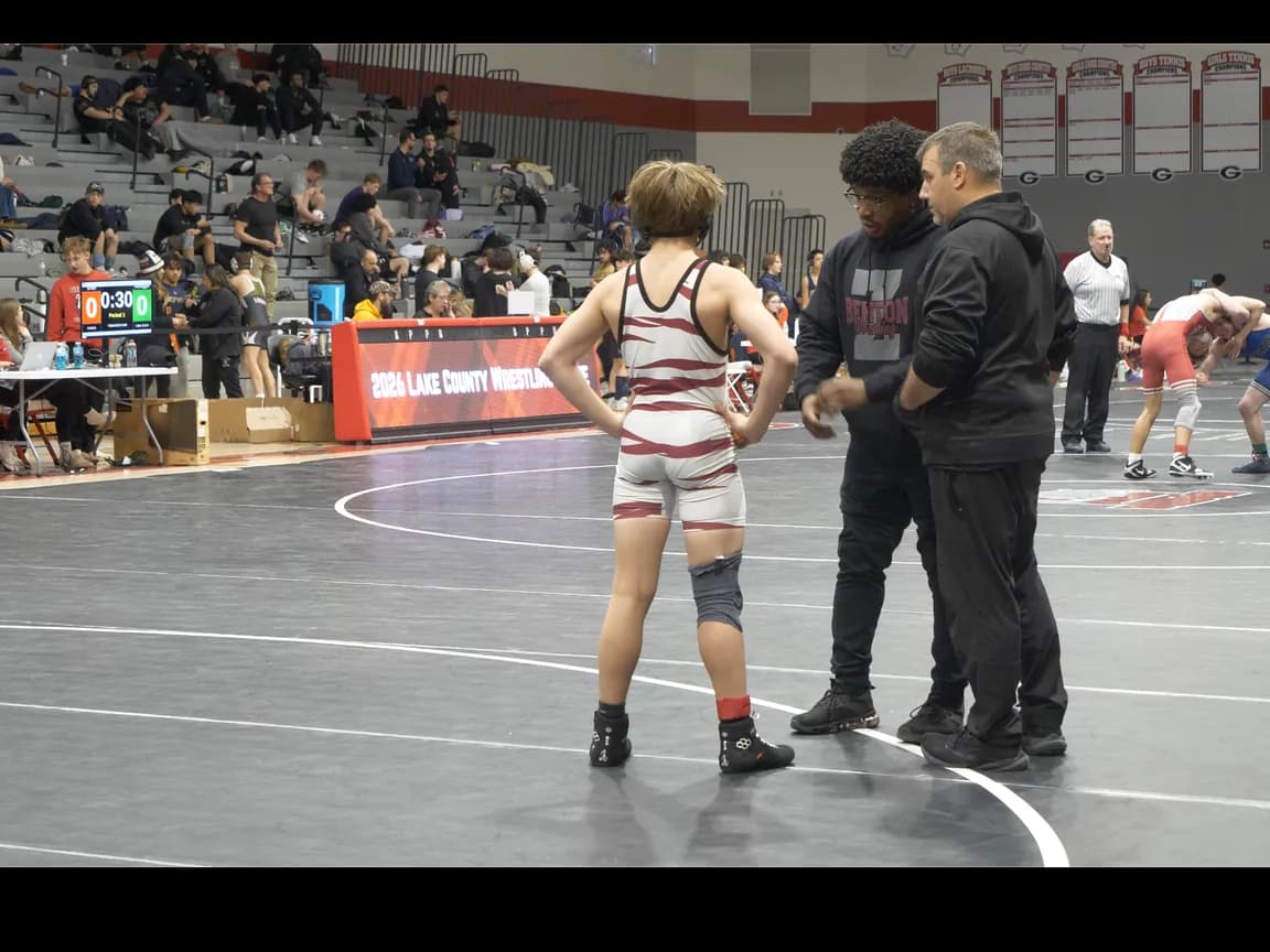 A coach giving between-match instruction to a singlet-wearing wrestler beside the mat at a tournament.